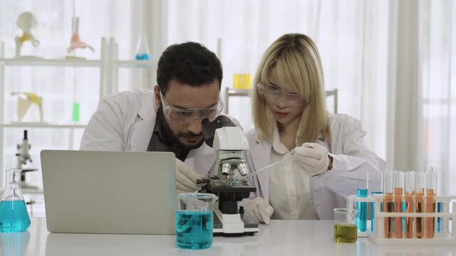 Two happy professional scientists and researchers in lab coats using a microscope and a laptop while conducting chemistry experiments with liquid in test tubes and beakers in the science laboratory. 