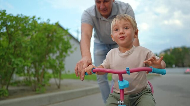 Happy father teaches little son to ride tricycle on paved path in summer park. Laughing boy sits on bike while dad helps him steer. Family enjoys outdoor fun. Caring parent pushes child on tricycle.