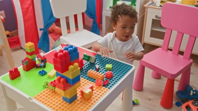 Little girl stands by a play table and reaches hand to stack colorful plastic building blocks with mouth open near pink chair in building; curiosity discovery learning.