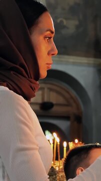 Woman in headscarf stands in church interior, side profile facing altar, with candles and ornate gold iconostasis behind her during prayer or worship, captured in reverent stillness.