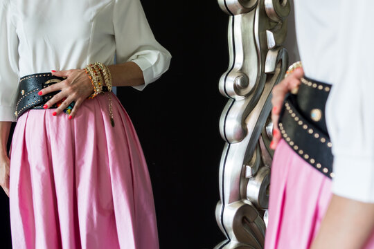 Cropped shot showing a woman wearing a white puff-sleeve top, a wide black studded belt, and a voluminous pink skirt, captured through a decorative silver mirror reflection.