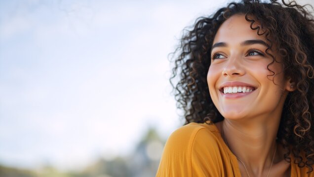 Close up portrait of a smiling woman with curly hair looking away. Happy young female face outdoors on a sunny day. Copy space for text