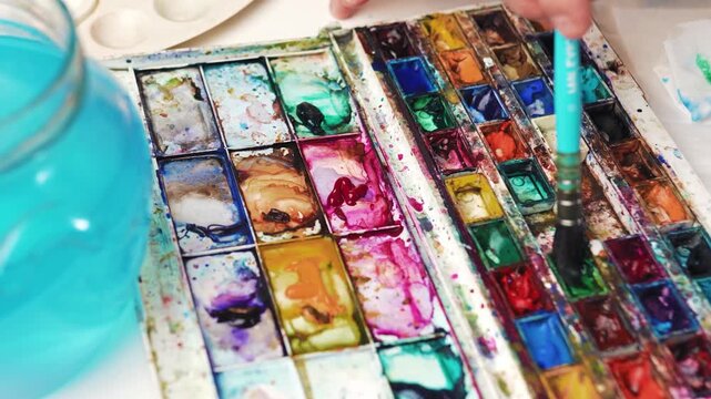 Closeup of artist hand dipping paintbrush into watercolor paint cuvette on palette