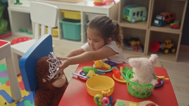 Little girl places a tiara on a plush monkey while hand reaches across a red play table in a building; playful childhood imagination wonder.