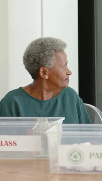Vertical video: Smiling at offscreen cue, senior woman in teal explaining GLASS or PAPER bins at tab