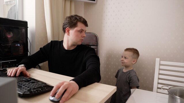 A young father tries to concentrate on his computer while his little son playfully distracts him. The boy seeks attention, showing the challenges of remote work and family life at home