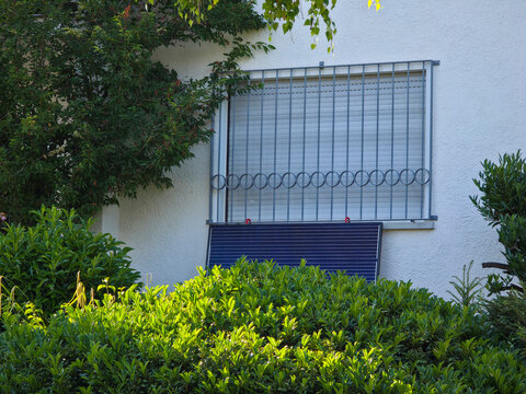 single pv module mounted beneath window along facade surrounded by lush green plants and blue sky
