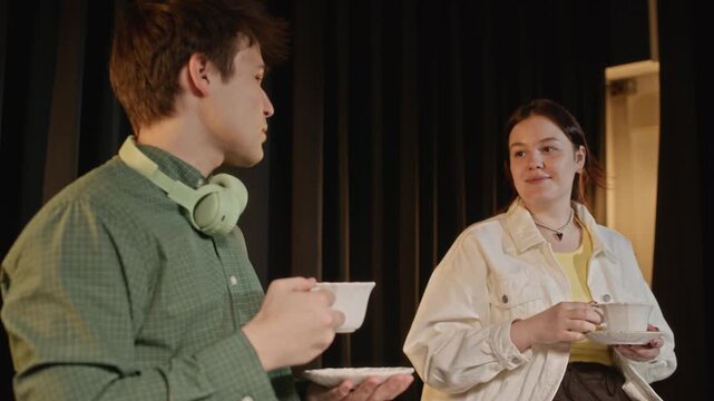 Panning shot of Caucasian teenage boy and girl rehearsing theatrical play on stage while holding teacups. For promoting youth drama clubs, acting classes, and performing arts education programs