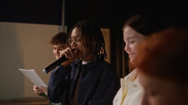 Dolly away shot of African American teenage boy speaking into microphone while rehearsing performance with classmates on stage. Ideal for educational marketing and youth talent development campaigns
