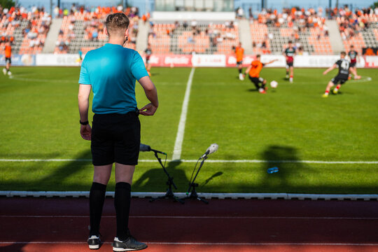 A soccer technical referee in a blue shirt stands on the sideline watching a football match in progress at a stadium.