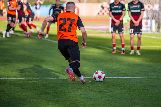 A soccer player in an orange jersey with number 27 runs with the ball toward the opponent's free kick wall during a match.
