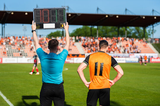 A soccer referee holding an electronic substitution board next to a player in an orange jersey waiting to enter the pitch.
