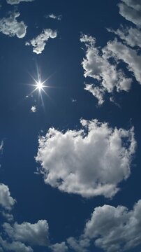 Vertical time lapse lookin straight up into a blue sky at the sun as clouds drift through the frame. A strong sun star is generated by the sun
