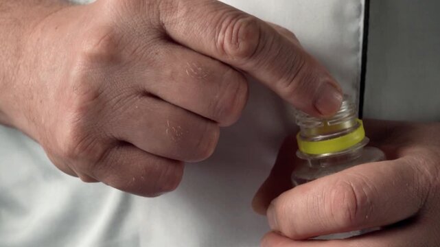 Hands of a person holding a soft gel capsule while preparing to open a bottle with a yellow seal in a well-lit indoor setting