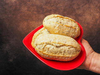 Naklejka premium Hand holds red heart shaped plate with three small sourdough rolls, grey painted background. High quality and value bread. Healthy bakery product. Love food concept. Ideal for food theme.
