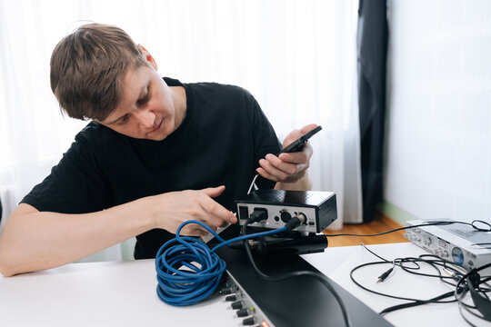 Focused man sets up audio equipment, connecting cables to sound interface with smartphone. Bright, natural lighting in modern home studio. Technical concept for music production or sound engineering.