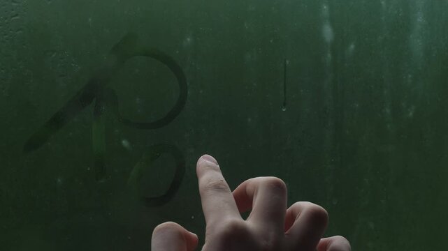 A person writes the word "Rain" with a finger on a foggy window glass during rain. Close-up of the inscription on the condensed glass with a blurred green background.