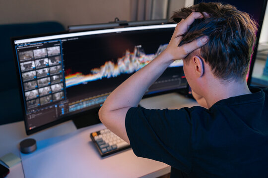 Young man in home office expressing frustration and stress while working on video editing software, color grading footage with timeline and waveforms visible on ultrawide monitor.
