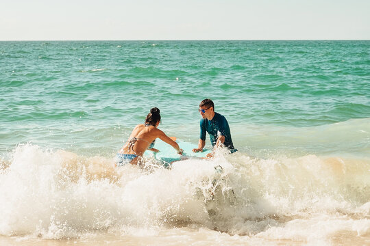 beach Ursa, Portugal.  Two friends sit on a surfboard while holding drinks as waves crash around them. Another person is seen paddling in background. sunny warm on beach.  waves crash  in warm weather