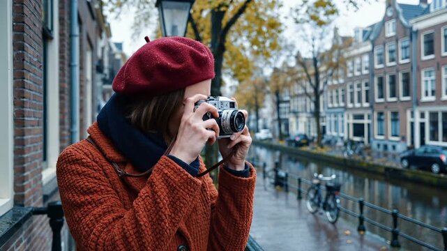 Young woman uses vintage camera near city canal. Female tourist wears red beret and coat. Photographer takes photo with camera. Woman holds camera and smiles. Tourist enjoys autumn canal view.