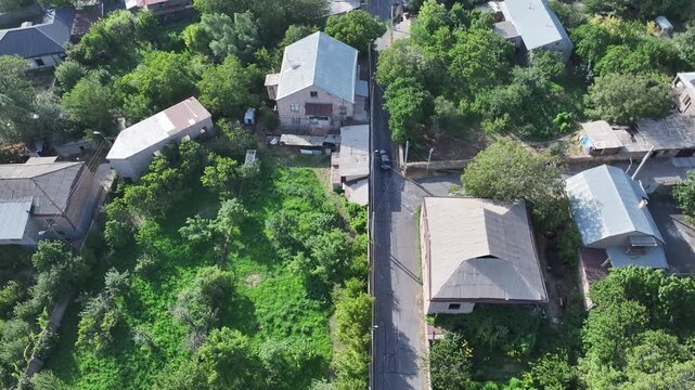 An aerial drone flight over a green suburban neighborhood with lush gardens, followed by a wide panoramic view of a sunny Armenian town surrounded by flat open fields.

