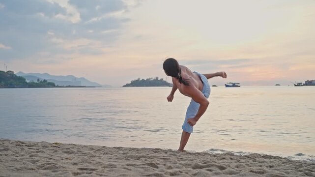Shirtless karate practitioner performing kicks on sandy beach at sunset training beside calm ocean with island on horizon warm sky, surf reflections, balanced athletic motion