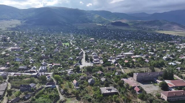 Beautiful aerial drone shot of the scenic Odzun village in Armenia, showing charming houses surrounded by lush green trees and majestic mountains under a bright summer blue sky.

