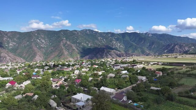 Beautiful aerial drone shot of the scenic Odzun village in Armenia, showing charming houses surrounded by lush green trees and majestic mountains under a bright summer blue sky.

