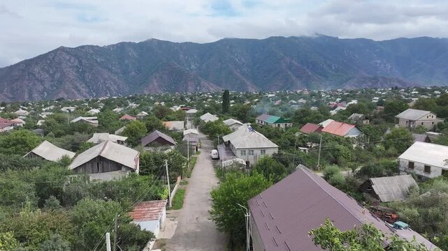 Beautiful aerial drone shot of the scenic Odzun village in Armenia, showing charming houses surrounded by lush green trees and majestic mountains under a bright summer blue sky.

