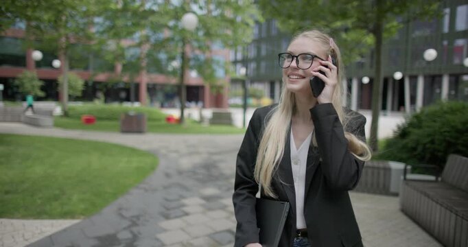 Young Caucasian female student wearing glasses in a modern urban environment among houses and trees walking in the park with a laptop and talking on the phone