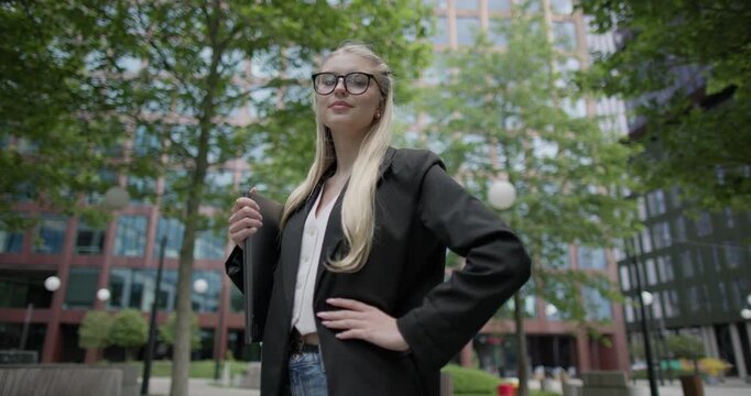 Young Caucasian female student wearing glasses in a modern urban environment among houses and trees standing in a park holding a laptop