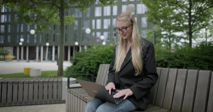 A young Caucasian female student wearing glasses in a modern urban environment among houses and trees sits on a bench in a park and works on a laptop