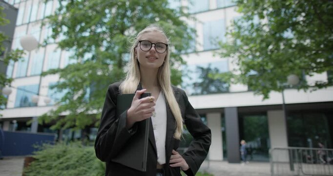 Young Caucasian female student wearing glasses in a modern urban environment among houses and trees standing in a park holding a laptop
