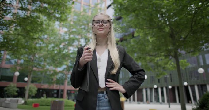 Young Caucasian female student wearing glasses in a modern urban environment among houses and trees standing in a park holding a laptop