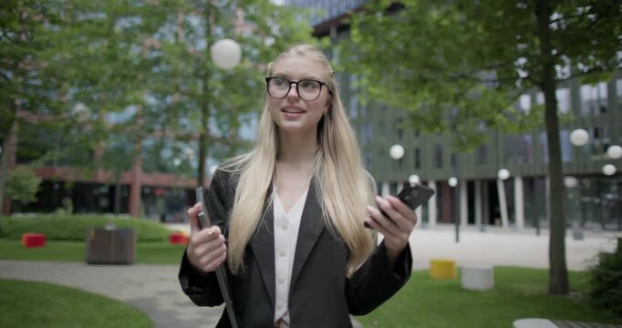 Young Caucasian female student wearing glasses in a modern urban environment among houses and trees walking in the park with a laptop and talking on the phone
