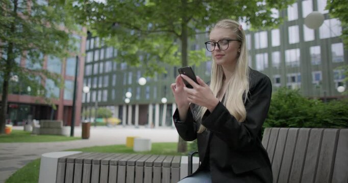 A young Caucasian female student wearing glasses in a modern urban environment among houses and trees sits on a bench in a park and uses a mobile phone - surfing the internet or social media