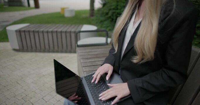 A young Caucasian female student wearing glasses in a modern urban environment among houses and trees sits on a bench in a park and works on a laptop
