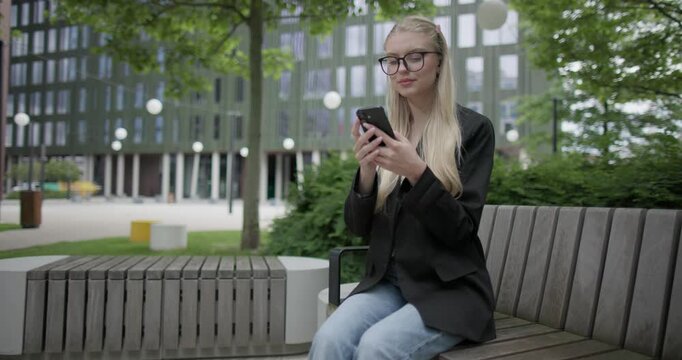 A young Caucasian female student wearing glasses in a modern urban environment among houses and trees sits on a bench in a park and uses a mobile phone - surfing the internet or social media