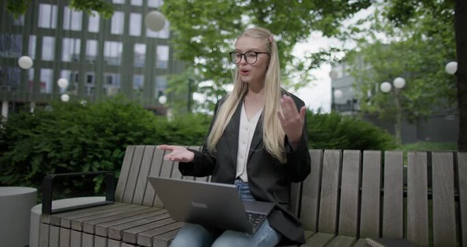 A young Caucasian female student wearing glasses in a modern urban environment among houses and trees sits on a bench in a park and works on a laptop conducting a video call, webinar or conference
