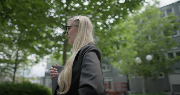 Young Caucasian female student wearing glasses in a modern urban environment among houses and trees standing in a park holding a laptop