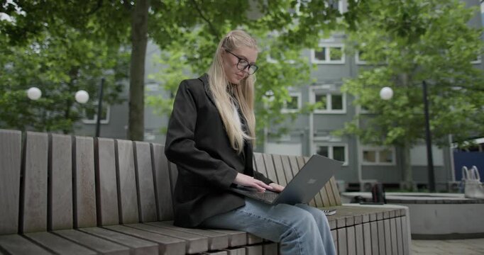A young Caucasian female student wearing glasses in a modern urban environment among houses and trees sits on a bench in a park and works on a laptop