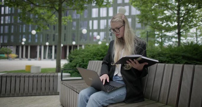 A young Caucasian female student wearing glasses in a modern urban environment among houses and trees sits on a bench in a park and works on a laptop reading notes from a notebook