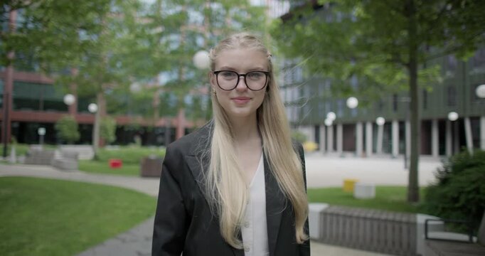A young Caucasian female student wearing glasses in a modern urban environment among houses and trees stands in a park and gestures with her hands