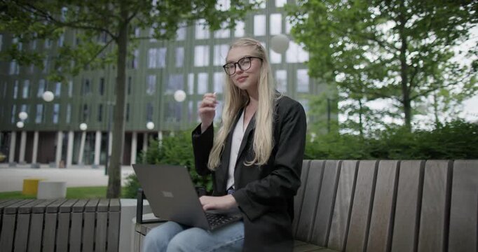 A young Caucasian female student wearing glasses in a modern urban environment among houses and trees sits on a bench in a park and works on a laptop