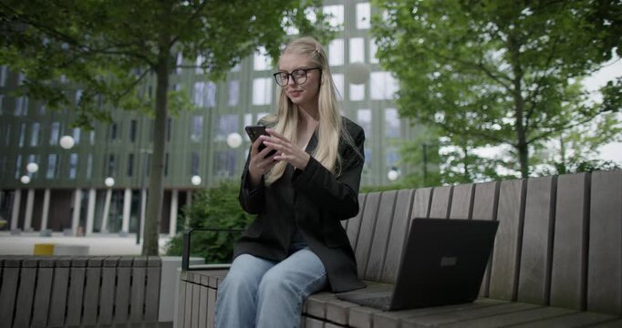 A young Caucasian female student wearing glasses in a modern urban environment among houses and trees sits on a bench in a park and works on a laptop and uses a mobile phone
