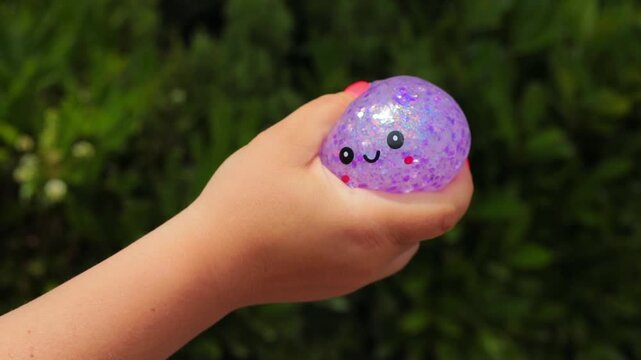 Hand squeezing purple glitter squishy dumpling toy with kawaii smiling face and red nail polish against green background