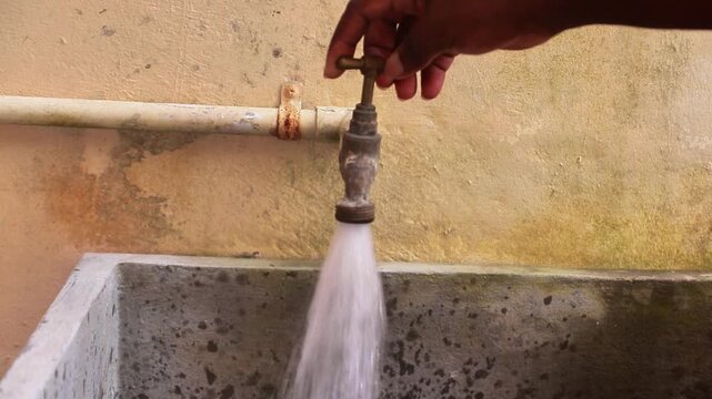 Running Water from Faucet: A hand turns on an old faucet, water streaming into a concrete basin, representing everyday utility and essential resources.