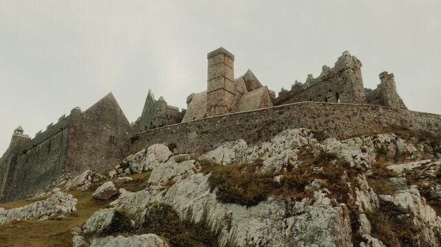 Aerial view of the Rock of Cashel featuring medieval stone ruins, a round tower, and ancient fortifications on a rocky hill under a cloudy sky in Cashel, County Tipperary, Ireland.