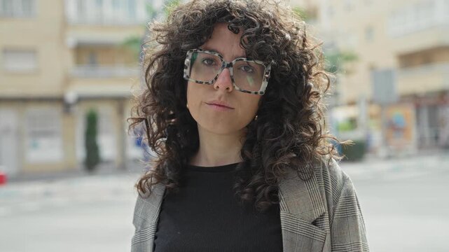 Woman with curly hair wearing oversized patterned glasses looks directly at camera on an urban street with buildings, storefronts and a parked car; thoughtful resilience.
