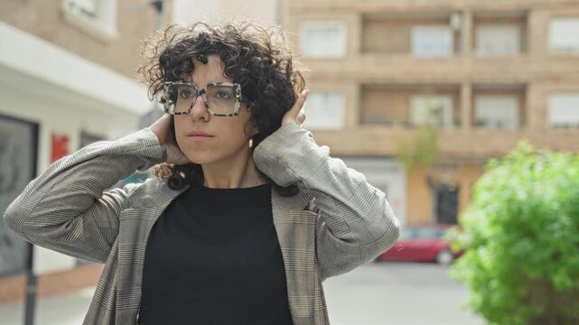 Woman adjusts oversized glasses and tucks curly hair on a street with apartment buildings, hands touching face and hair; quiet confidence.